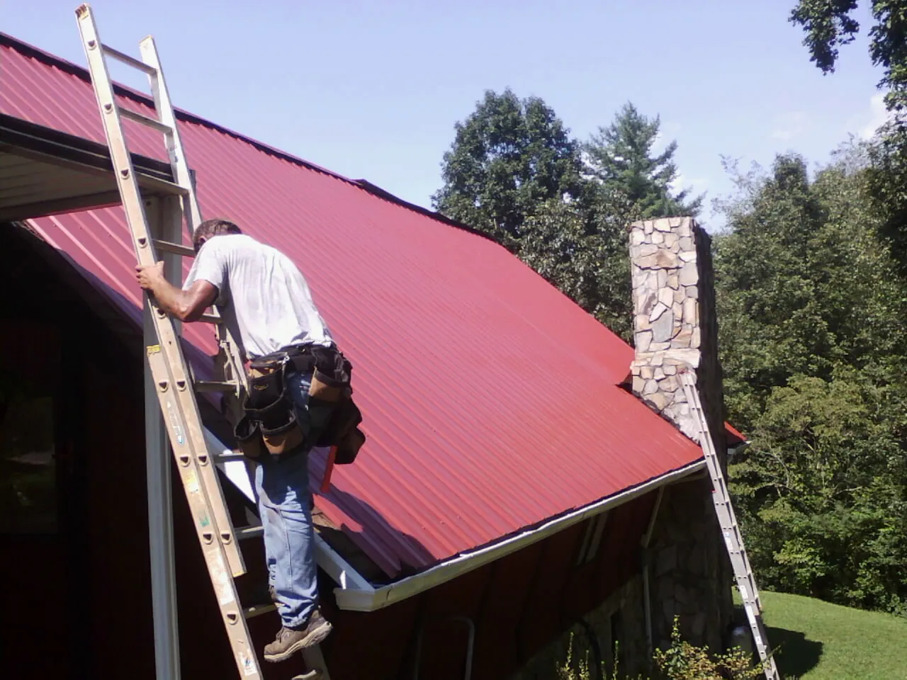Roofer on a ladder beside a red metal roof and stone chimney — Soffit & Fascia Repair by Roofing Brothers in Upper Marlboro